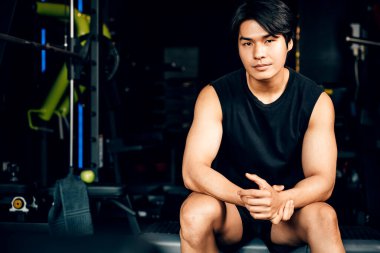 Asyalı genç adam portresi siyah spor kıyafetleri içinde dinlenmek için oturmuş spor salonunda su ya da protein tutarken ağırlık antrenmanı, sağlıklı yaşam tarzı konsepti.