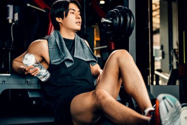 Spor kıyafetleriyle eğitilmiş Asyalı bir gencin portresi spor salonunda antrenman, ağırlık eğitimi, sağlıklı yaşam tarzı konsepti sonrasında elinde su ya da proteinle bankta dinleniyor.