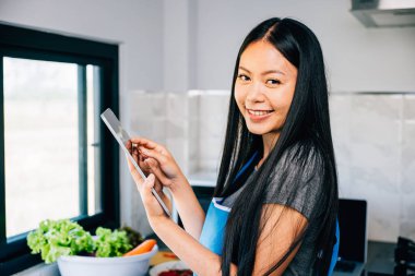 Güneşli bir mutfakta gülümseyen Asyalı bir kadın, teknolojinin ve mutfak sanatının neşeli bir karışımını sunarken tabletle etkileşime geçiyor..