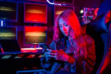 Bilgisayar ekranının önündeki Asyalı kadın cep telefonu, neon ışık, Hacker sistem kontrol odasında siyah ceket giyiyor güvenlik görevlisi birden fazla monitörde birlikte çalışıyor akıllı telefon.