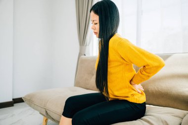 Dayanılmaz sırt ağrısı çeken, koltukta oturan Asyalı kadın belini tutuyor. Kronik sırt ağrısı rahatsızlığı ve sırt sorunları için tıbbi bakım gerekliliği. sağlık hizmeti kavramı