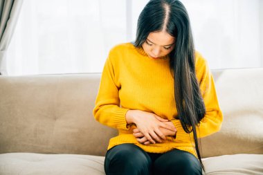 Karın ağrısı çeken, koltukta oturan, şiddetli karın ağrısından muzdarip genç bir kadın. Rahatsızlık belirtilerini vurgulamak ve karın sağlığına dikkat çekmek. Sağlık hizmeti karın şişkinliği konsepti