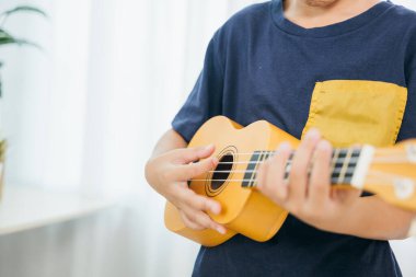 Mavi tişörtlü genç bir çocuğun oturma odasında sarı ukulele çalarken yakın plan fotoğrafı. Arka plandaki yumuşak beyaz perdeler sahne, müzik ve yaratıcılığa olan tutkusunu yakalıyor..