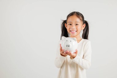 Asyalı küçük bir kız elinde domuz kumbarası, beyaz arka planda izole edilmiş stüdyoya bakıyor, mutlu bir anaokulu çocuk yaşam tarzı gülümsüyor, beyaz kumbara, Tayland kişisel para tasarrufu.