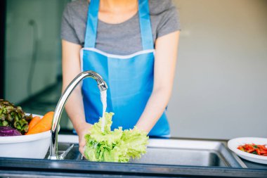 Ev mutfağında yemek pişirmekten zevk alan gülümseyen bir kadın lavaboda taze sebzeleri yıkıyor ve lezzetli bir salata için hazırlıyor. Sağlıklı bir yaz yemeği için açık yeşil yapraklar..