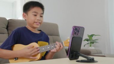 Akıllı telefon kullanarak müzik dersini canlı yayınlarken ukulele çalan Asyalı çocuk. Monte edilmiş telefon standı ve rahat oturma odası sahnesi modern öğrenme ve evde yaratıcı faaliyetler sergiliyor..