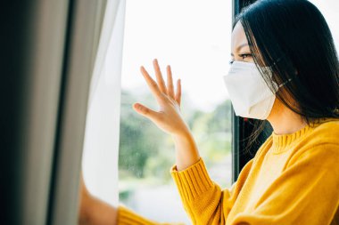 COVID 19 izolasyonunu yansıtan koruyucu maskeli kadın üzgün ve yalnız hissederek pencereden dışarı bakıyor. Korku yalnızlığını ve salgın krizinin etkilerini yansıtıyor..
