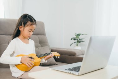 Kanepede oturan kız çocuğu dizüstü bilgisayar videosundan rehberlik alarak ukulele çalışıyor. Huzurlu bir oturma odası evde teknoloji ve müzik eğitiminin entegrasyonunu gösteriyor.