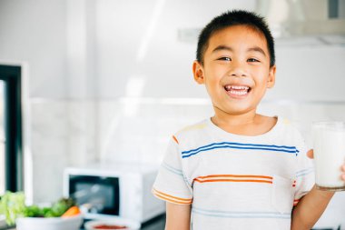 Neşeli Asyalı küçük çocuk mutfakta süt tutuyor, mutlu bir şekilde gülümsüyor. İçkiden zevk alan tatlı bir oğlun portresi. Neşeli çocuk kalsiyum zengini sıvıyı yudumluyor, evde mutluluk saçıyor..