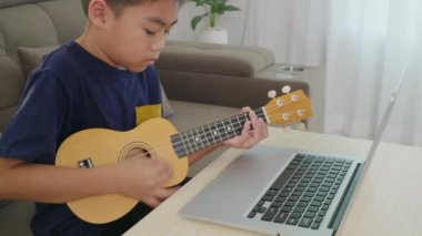 Çocuk kanepede ukulele öğrenirken dizüstü bilgisayarda özel ders izliyor. Konforlu oturma odası sahnesi, teknolojinin ve yaratıcı eğitimin modern ev ortamında entegrasyonunu vurguluyor..