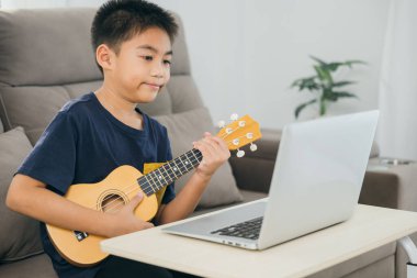 Genç bir çocuk, oturma odasında bir kanepede ukulele ile oturuyor ve dizüstü bilgisayarından online bir ders izliyor. Çalmayı öğreniyor, modern teknolojiyle müzik pratiğini birleştirerek rahat bir ev ortamında..
