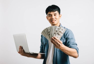 Asyalı gülümseyen genç adam dizüstü bilgisayar ve para fanatiği stüdyo görüntüsü izole beyaz arka plan, mutlu yaşam tarzı işadamı dolar banknotları içinde para tutuyor.