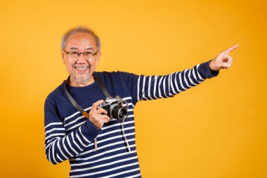 Asyalı, gözlüklü yaşlı bir adam klasik SLR kamera tutuyor ve stüdyo tarafından çekilmiş bir şeyi işaret ediyor izole edilmiş sarı arka plan, gülümseyen yaşlı fotoğrafçı gri saçlı yaşlı adam.