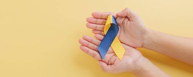 Down Sendromu Günü. Sarı ve mavi farkındalık kurdelesi nazikçe sarı arka planda elle tutulur. Down Sendromu ve Otizm Bilinci İçin Küresel Çabaları Destekleme.