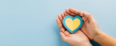 Down Sendromu ve Otizm Bilinci küresel düzeyde. Mavi kenarlı sarı bir kalp, Down Sendromu farkındalığı için sarı arka planda olan eller tarafından tutulur..