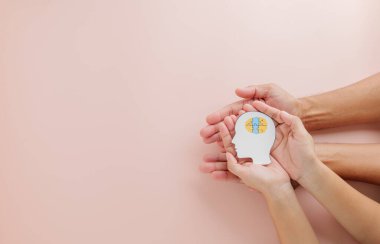 Yetişkin ve çocuk elleri Down Sendromu farkındalığı için kesilmiş bir kağıt parçasında beyin yapbozu tutuyor. Down Sendromu Günü 'nü ve Otizm Farkındalığını pembe arka planda küresel bir umut mesajıyla kutluyoruz..