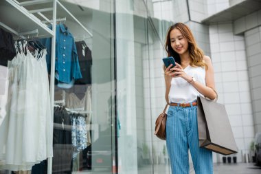 Moda mağazasının önünde akıllı telefonunu kullanan genç bir kadın internetteki alışveriş ve moda penceresine gülümsüyor. Çantaları en son giysi ve aksesuarlarla dolu..