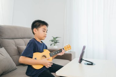 Kanepede ukulele çalan bir çocuk, canlı yayın veya internet dersleri için öne monte edilmiş bir akıllı telefon. Sahne, geleneksel müzikle ev ortamında yenilikçi dijital öğrenmeyi harmanlıyor..