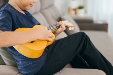 Kanepede oturan, sarı bir ukulele çalan genç bir müzisyenin yakın çekimi. Mavi gömlek ve sıcak ortam neşeli bir müzik anını vurguluyor..