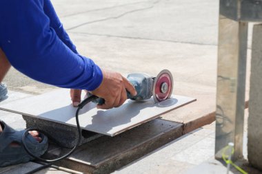 Mavi elbiseli bir işçi bir taş parçasındaki gri fayansı kesmek için bir açı öğütücü kullanıyor. Yakından bakıldığında, bir inşaat alanındaki fayans kesiminin ustalığı ve hassasiyeti vurgulanıyor..