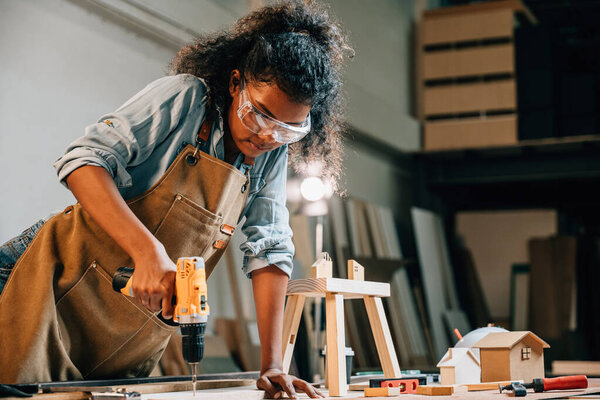 Плотница, использующая дрель, чтобы закрепить кусок дерева в мастерской, фокусируется на точности в производстве мебели. Окруженный инструментами и деревянными деталями, подчеркивая мастерство и мастерство
