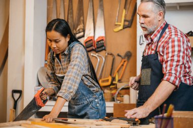 Kıdemli marangoz, atölyede odun kesmek için el testeresi kullanan bir kadına rehberlik eder. Aletler ve ahşap malzemelerle çevrili iyi donanımlı bir yerde ahşap işçiliği projesinde birlikte çalışıyorlar.