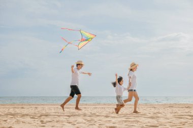 Asyalı bir aile sahilde koşarken gökyüzünde uçurtma uçuruyor. Mutlu ebeveynler, anne ve babalar ve çocukları uçurtmayla oynuyorlar, aileler birlikte vakit geçiriyorlar, yaz günü.