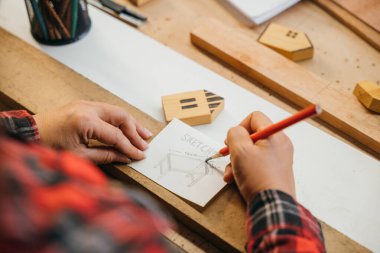 Kırmızı ekose gömlekli biri kırmızı kalemle çizim masası planları yapıyor. Tahta tahtalar ve minyatür evlerle çevrili. Mimarlık, inşaat ve DIY proje kavramları için mükemmel