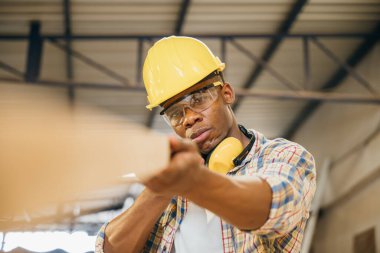 Afro-Amerikan marangoz atölyede tahta taşırken bir tahta parçasını teftiş ediyor. İnşaat işleri, ahşap kalite teftişi ve marangozluk, Ulusal Marangozlar Günü.
