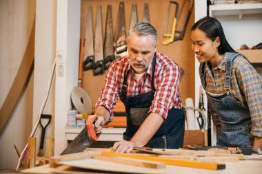 Kıdemli marangoz, atölyede odun kesmek için el testeresi kullanan bir kadına rehberlik eder. Aletler ve ahşap malzemelerle çevrili iyi donanımlı bir yerde ahşap işçiliği projesinde birlikte çalışıyorlar.