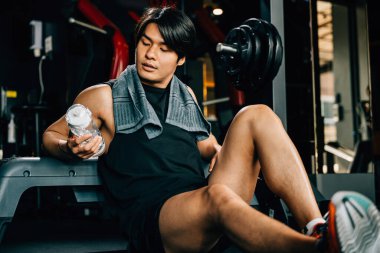 Spor kıyafetleriyle eğitilmiş Asyalı bir gencin portresi spor salonunda antrenman, ağırlık eğitimi, sağlıklı yaşam tarzı konsepti sonrasında elinde su ya da proteinle bankta dinleniyor.