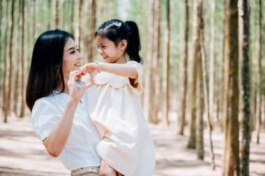 Huzurlu bir ormanda kızını kucağında taşıyan Asyalı bir anne, mutlu bir gülümseme anne ve küçük çocuk, elleriyle kalp işareti yapıyor birlikte hassas anın tadını çıkarın, aile günü anısı tatil konseptinde.