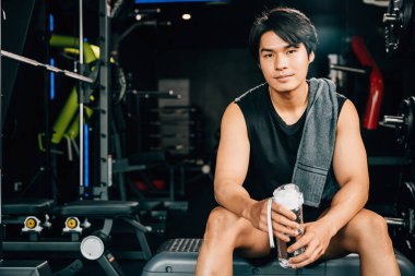 Asyalı genç adam portresi siyah spor kıyafetleri içinde dinlenmek için oturmuş spor salonunda su ya da protein tutarken ağırlık antrenmanı, sağlıklı yaşam tarzı konsepti.