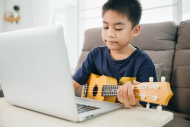 Ukulele çalışan bir çocuk, elleri odaklı, online ders gösteren bir dizüstü bilgisayarla. Yakın çekim, teknoloji ve geleneksel müziğin bir ev ortamında entegrasyonunu vurguluyor.