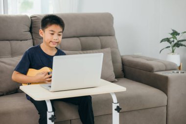 Evde ukulele çalışması yapan genç bir çocuk, bir dizüstü bilgisayarla çevrimiçi kursu takip ediyor. Oturma odasındaki kanepe dijital eğitim ve kendini ifade etme yoluyla müzik öğrenmek için rahat bir alan yaratıyor..