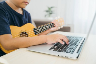 Dizüstü bilgisayardan özel ders izlerken ukulele çalan genç bir çocuğun yakın plan fotoğrafları. Oturma odasındaki kanepede oturarak geleneksel müzik pratiğini modern öğrenme araçlarıyla harmanlıyor..