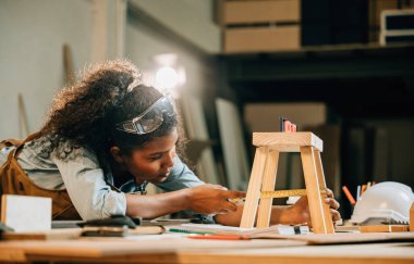 Afro-Amerikalı marangoz kadın bir dizüstü bilgisayar kullanarak ahşap bir projede çalışıyor. Yeni mobilyalar tasarlamak için modern teknolojiyi kullanın, el işçiliğini dijital aletlerle harmanlayın, Ulusal Marangozlar Günü
