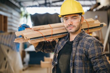 Sarı kasketli ve ekose gömlekli gülümseyen inşaat işçisi omzunda tahta tutuyordu. Marangozluk, marangozluk ve mobilya atölyesinde işçilik için ideal, Ulusal Marangozlar Günü