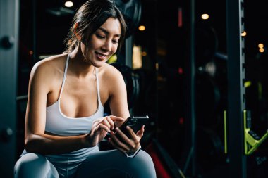 Asyalı kadın yedek kulübesinde dinleniyor ve beden eğitimi aldıktan sonra tüm güncellemeleri kontrol ediyor. Kadın spor salonunda spor yaparken cep telefonu uygulaması kullanıyor. Sağlıklı yaşam tarzı konsepti.