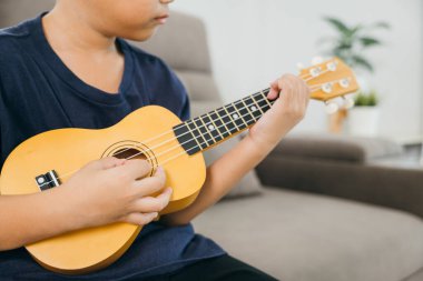Parlak gülüşlü genç çocuk gri bir kanepede ukulele çalıyor. Konforlu oturma odası ve enstrümana odaklanma mükemmel bir neşe, rahatlama ve sanatsal ifade karışımı yaratır..