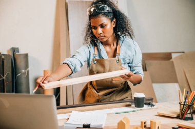 Marangoz kadın bir atölyede bir odun parçasını tutuyor ve inceliyor. Alet ve malzemelerle çevrili, marangozluk ve işçilik üzerine odaklanmış, Ulusal Marangozlar Günü
