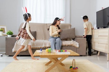 Paskalya kutlaması. Tavşan kulaklı bir anne ve çocuklar, Paskalya yumurtalarını oturma odasında ararken kahkaha ve neşeyi paylaşırlar. Aile eğlencesi.