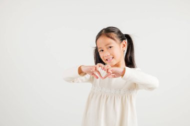 Gülümseyen Asyalı genç bir kız güven gösteriyor. Beyaz arka planda elleri ve parmakları olan bir kalp şekli yaratıyor. Anaokulu çocuğunun portresi Sevgililer Günü 'ne sevgilerini gönderiyor.