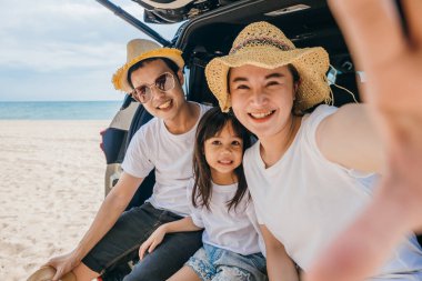 Mutlu Asyalı aile, sahilde park etmiş beyaz bir cipin arkasında otururken akıllı telefonuyla selfie çekerken eğleniyor. Yaz gezisi konsepti.
