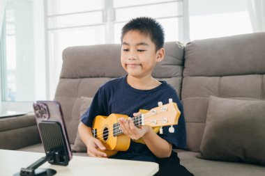Kanepede oturan genç bir çocuk, canlı müzik dersleri için akıllı bir telefonla uğraşırken ukulele çalıyor. Kurulum, teknoloji meraklısı, yaratıcı ev temelli öğrenmenin özünü yakalıyor..