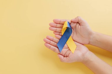 İki el nazikçe sarı arka planda Down Sendromu Günü 'nü sembolize eden sarı ve mavi farkındalık kurdelesini tutuyor. Dünya çapında Down Sendromu ve Otizm Bilinci Tanıtımı.
