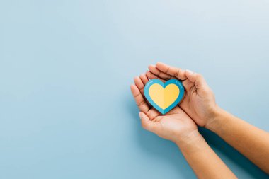 Eller, içinde sarı bir kalp ve Down Sendromu farkındalığı için sarı arka planda açık mavi bir kenar tutuyor. Küresel Down Sendromu ve Otizm Farkındalığı Çabalarını Destekleme.