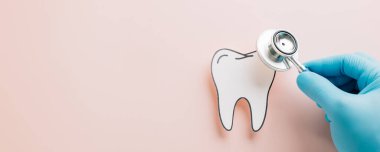 Eldivenli bir doktorla dişçi günü. Pembe arka plandaki kağıt dişleri kontrol etmek için steteskop kullanıyor. Diş bakımı ve ağız temizliğini kutluyoruz..