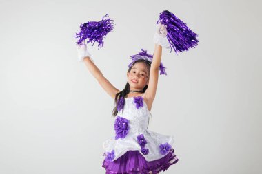 Ponpon kız üniforması içindeki heyecanlı çocuk mor ponponları tutuyor. Beyaz arka planda stüdyo çekimi, neşe, eğlence ve neşe dolu. Amigo kız.