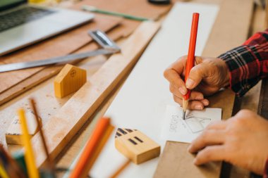 Kırmızı ekose gömlekli biri kırmızı kalemle çizim masası planları yapıyor. Tahta tahtalar ve minyatür evlerle çevrili. Mimarlık, inşaat ve DIY proje kavramları için mükemmel